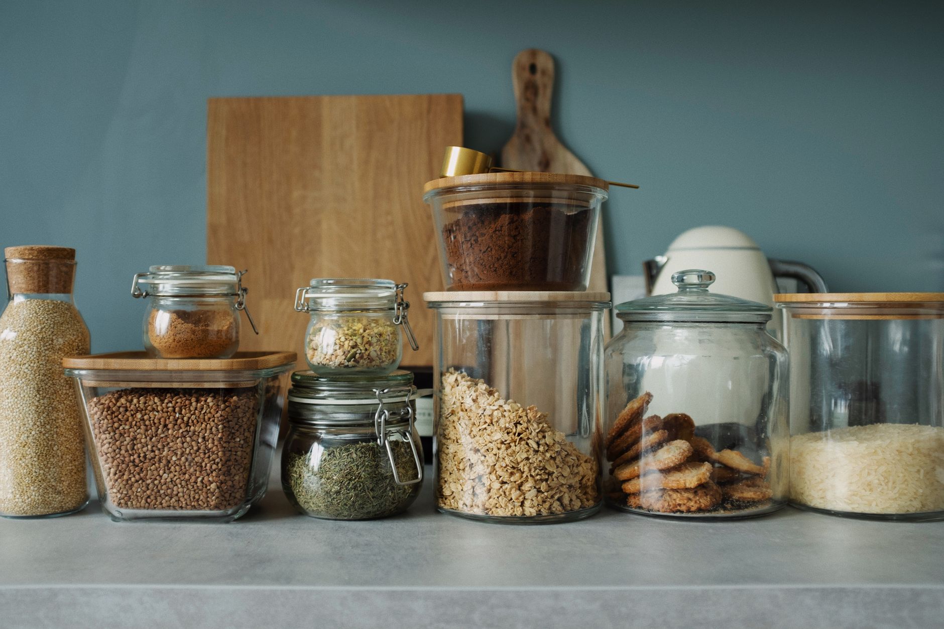 Bocaux et boîtes en verre alignés sur un plan de travail de cuisine, remplis de flocons, de riz, de graines, d’herbes et de biscuits.