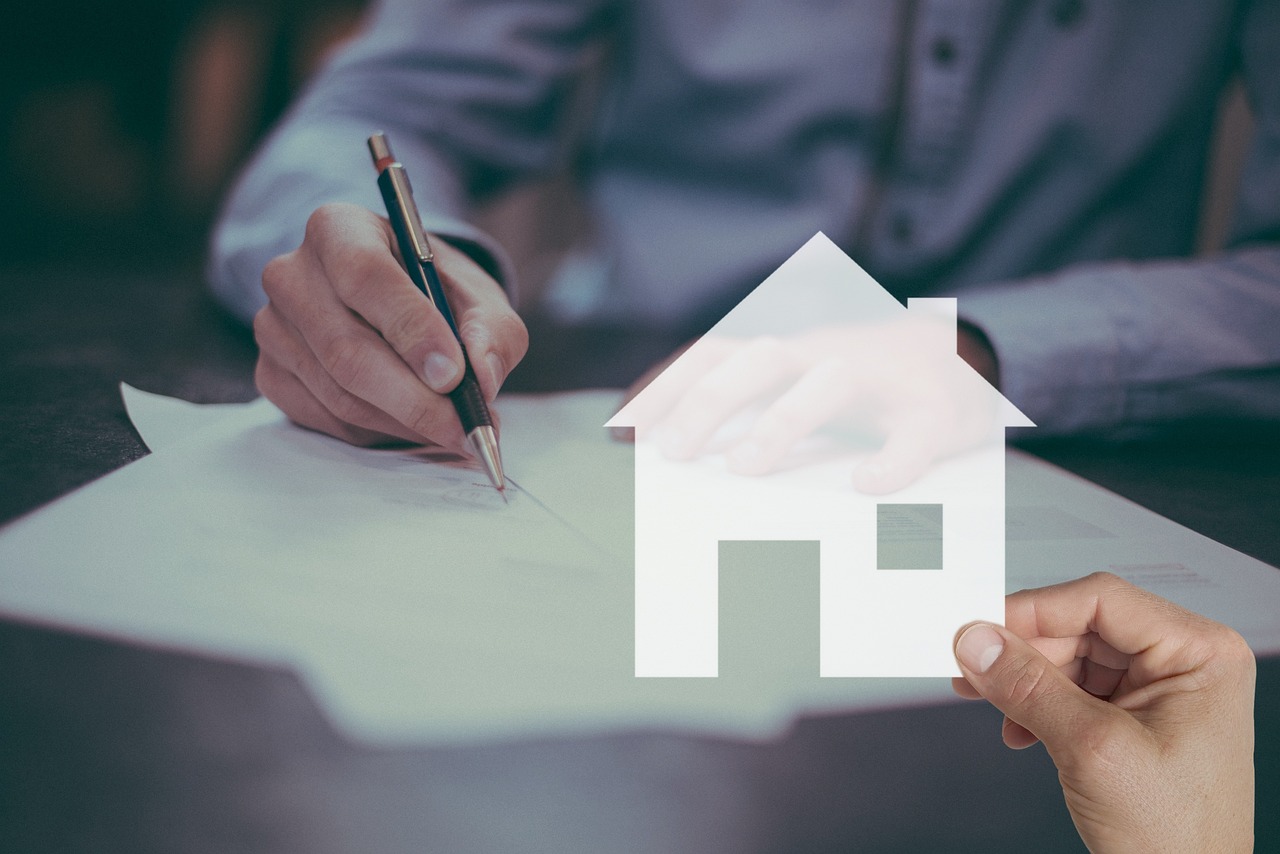 Mains d'une personne qui signe un document avec un stylo, devant une silhouette blanche de maison découpée.